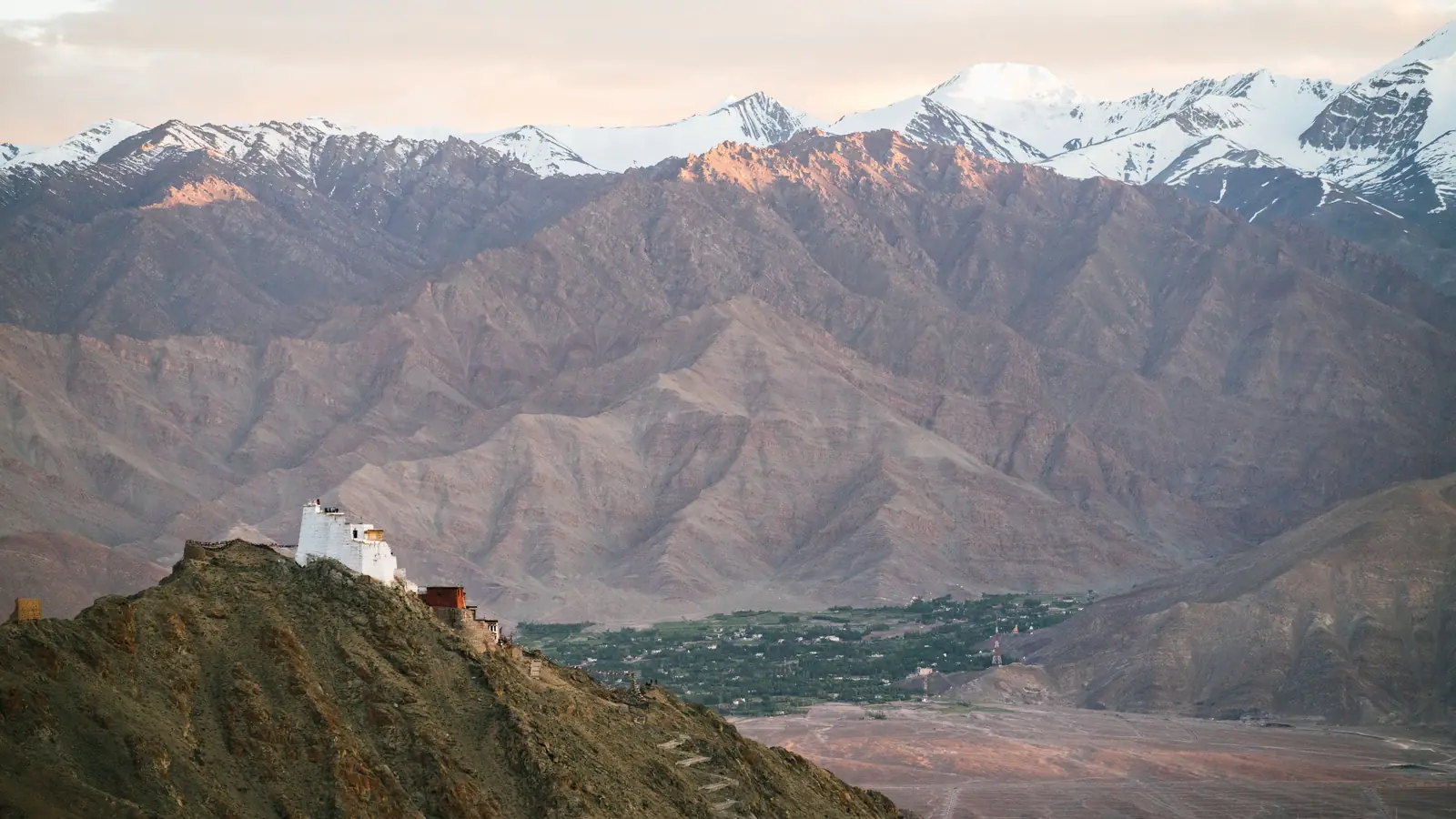 A white monastery on a bare ridge above a green valley, with snow peaks behind, in Ladakh